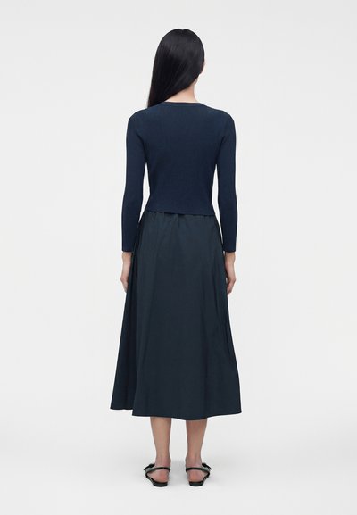 Femme aux cheveux longs, raides et noirs, portant un haut à manches longues bleu marine, une jupe midi et des sandales plates noires, debout de dos sur fond blanc.