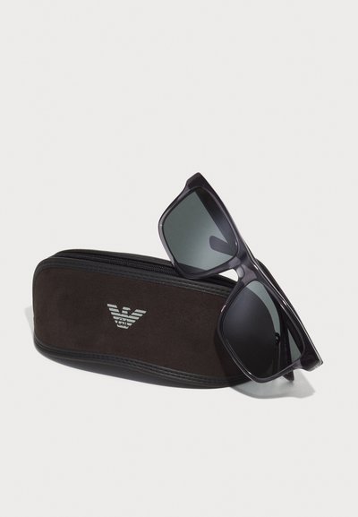 Ochelari de soare negri cu un cadru pătrat, lentile gri, așezați pe o carcasă din suede de culoare brun închis, cu un logo argintiu. Textură netedă și închidere cu fermoar.