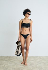 Μαύρο σετ μπικίνι με εφαρμοστό μπραλέτα και χαμηλής μέσης σλιπ, με οριζόντιες ραβδώσεις. Το μοντέλο κρατά μια τσάντα με μαύρες και καφέ ρίγες.