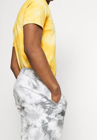 Person med gul batik-T-shirt och grå batik-mjukisbyxor med händerna i fickorna, visad från sidan mot en vit bakgrund.