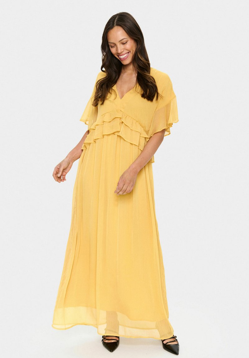 Yellow maxi dress with layered ruffle accents, V-neckline, and flowing fabric. Sheer texture and pleats, paired with black pointed-toe shoes.