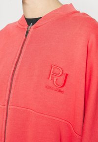 Sudadera con cremallera en color coral, hecha de un tejido suave. Presenta un logotipo en relieve "PJ" en el pecho, una cremallera en contraste y puños acanalados.