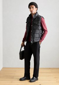 Čierny puffový vest s patentkami, nosený cez vínový sveter. Čierne nohavice a mokasíny dopĺňajú outfit, ktorý je doplnený čiernou taškou.