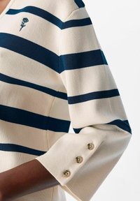 Gestreifter Pullover in Creme und Marineblau, mit V-Ausschnitt, Dreiviertelärmeln und dekorativen Knöpfen an den Bündchen sowie einem Logodetail.