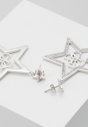 Boucles d'oreilles en argent en forme d'étoile avec des cristaux incrustés et un design décoratif de lettres entrelacées au centre, reposant sur une surface blanche.