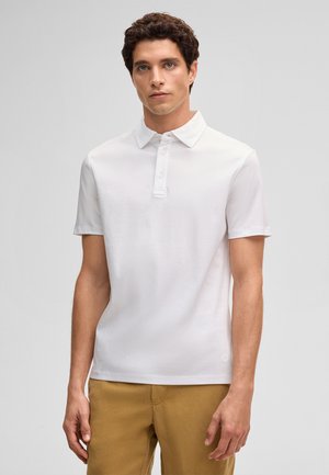 Młody mężczyzna w białej koszulce polo z krótkim rękawem i musztardowych spodniach na jednolitym szarym tle.