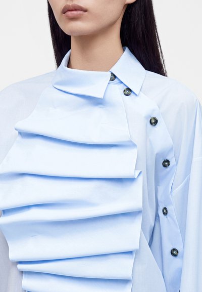 Femme portant une chemise bleu clair avec un devant plissé asymétrique et des boutons foncés le long de l'épaule et de la poitrine.