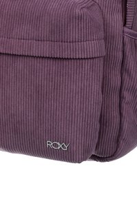 Paarse corduroy tas met een ribbelstructuur, voorzien van een voorvak en een zilveren Roxy-logo. Compacte vorm met een zachte afwerking.