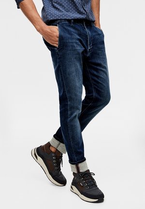 Homme portant un jean bleu foncé retroussé, une chemise bleue à motifs et des baskets noires et marron, debout les mains dans les poches.