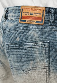 Πίσω τσέπη από ανοιχτό μπλε ξεθωριασμένο τζιν με δερμάτινη ετικέτα μάρκας Diesel Industry σε μπεζ και μπλε χρώμα πάνω από την τσέπη.