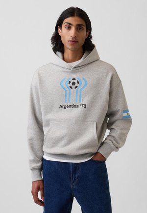 Tânăr cu păr închis la culoare, purtând o hanorac gri cu un design cu minge de fotbal albastră și neagră și textul „Argentina '78”, asortat cu blugi albaștri.