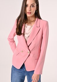 Rosa doppelt knopfbarer Blazer mit goldenen Knöpfen, Reverskragen und seitlichen Taschen, kombiniert mit einer hellen Bluse mit floralem Muster und Jeans.