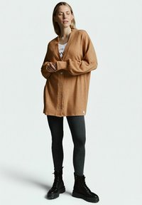 Brauner, übergroßer Cardigan aus weichem Stoff, mit tiefem V-Ausschnitt, großen Knöpfen und langen Ärmeln, kombiniert mit schwarzen Leggings und Stiefeln.