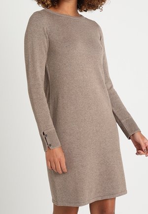 Kobieta ubrana w taupe'ową dzianinową sukienkę do kolan z długim rękawem, z prążkowanymi mankietami i detalami z guzikami na rękawach.