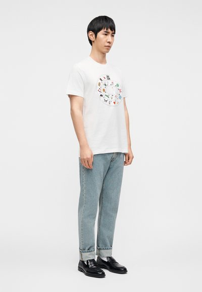 Λευκό T-shirt με κοντά μανίκια που έχει έναν κυκλικό σχεδιασμό με πολύχρωμες απεικονίσεις ζώων. Φορεμένο με τζιν ανοιχτού χρώματος και μαύρα παπούτσια.