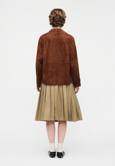 Άτομο με κοντά μαλλιά που φοράει καφέ σανούα σακάκι, μπεζ ριγωτή φούστα, λευκές κάλτσες και μαύρα παπούτσια, στέκεται γυρισμένο από πίσω.