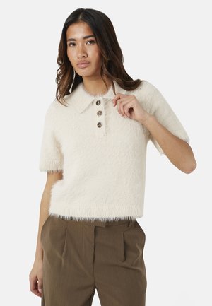 Donna con i capelli lunghi che indossa un maglione corto crema e morbido con bottoni e pantaloni marroni a vita alta su sfondo bianco.