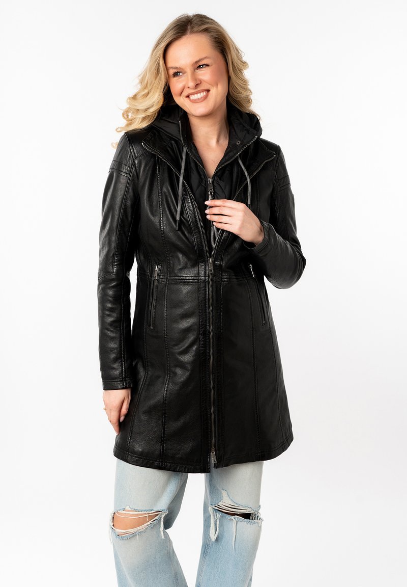 Zwarte leren lange jas met een ritssluiting, capuchon en voorzakken; heeft een gladde textuur en een getailleerd ontwerp. Draagt over versleten jeans.
