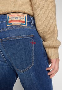 Blå denimjeans har ett lädermärke med texten "Diesel Industry" på baksidan. De har en röd logostickning på fickan.