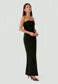 Melnā strapless kleita ar pieguļošu siluetu, ar drapētiem elementiem pie krūšu zonas un garumu līdz grīdai. Valkāta ar aso purngalu kurpēm.