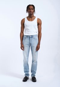 Jeans de color azul claro con un corte recto, cinco bolsillos y un cierre clásico de botones. Combinados con una camiseta sin mangas blanca y zapatos negros.