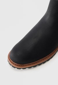 Punta de bota de cuero negro texturizado con ala cosida marrón y suela de taco negro sobre un fondo blanco.
