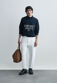 Marinblå sweatshirt med vitt "Emporio Armani"-logotyp, vita skräddarsydda byxor, svarta sneakers och brun läderväska. Ren, modern design.