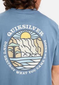 Osoba v modrém tričku Quiksilver s kulatým přírodním vzorem zobrazujícím vodopád, slunce, stromy a nápis "Nenič to, co jsi přišel užívat."