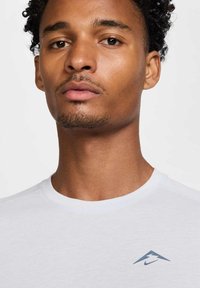 Camiseta blanca de manga corta con cuello redondo y un pequeño logo triangular azul en la parte izquierda del pecho. Textura de tela suave visible.