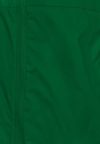 Groen nylonstof met een gladde textuur en zichtbare stiksels. Heeft een centrale rits die niet volledig geopend is en opgaat in de stof.