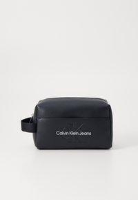 Crna pravokutna kozmetička torbica izrađeno od glatkog materijala, s zatvaranjem na patentni zatvarač i ugraviranim logotipom "Calvin Klein Jeans" sa strane.