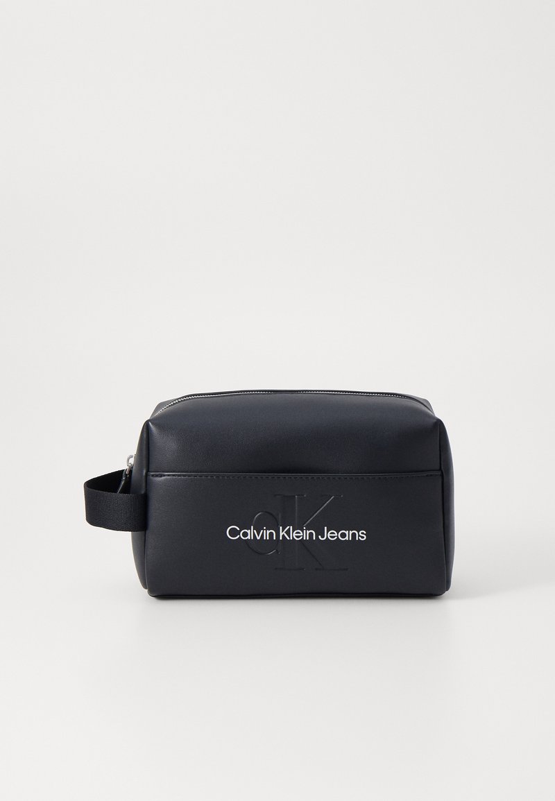 Crna pravokutna kozmetička torbica izrađeno od glatkog materijala, s zatvaranjem na patentni zatvarač i ugraviranim logotipom "Calvin Klein Jeans" sa strane.