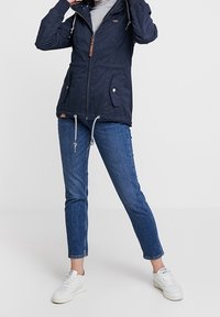 Navy waterproof jacket with a zip front, drawstrings, two side pockets, and a hood. Paired with blue slim jeans and white sneakers.