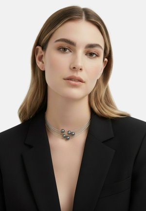 Metaliczny naszyjnik obroża z trzema paskami i pięcioma ciemnymi koralikami o gładkiej fakturze. Noszony na czarnym żakiecie, prezentujący dopasowany krój.
