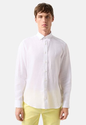 Mand iført en hvid skjorte med knapper og lange ærmer samt lysegule bukser, stående mod en ensartet lys baggrund.