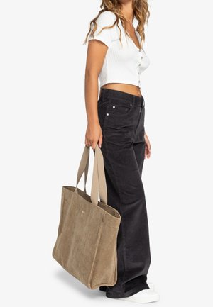 Femme portant un haut blanc court à boutons et un pantalon noir, portant un grand sac fourre-tout beige texturé avec de larges poignées.