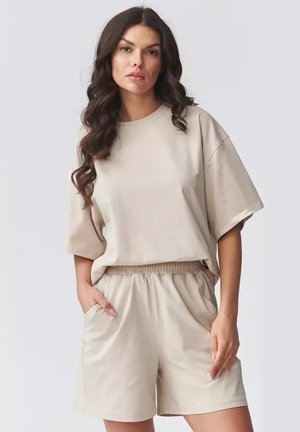 Kobieta o długich, falowanych włosach, ubrana w beżowy, oversize'owy t-shirt i dopasowane szorty z elastycznym pasem, ręce w kieszeniach, neutralne tło.