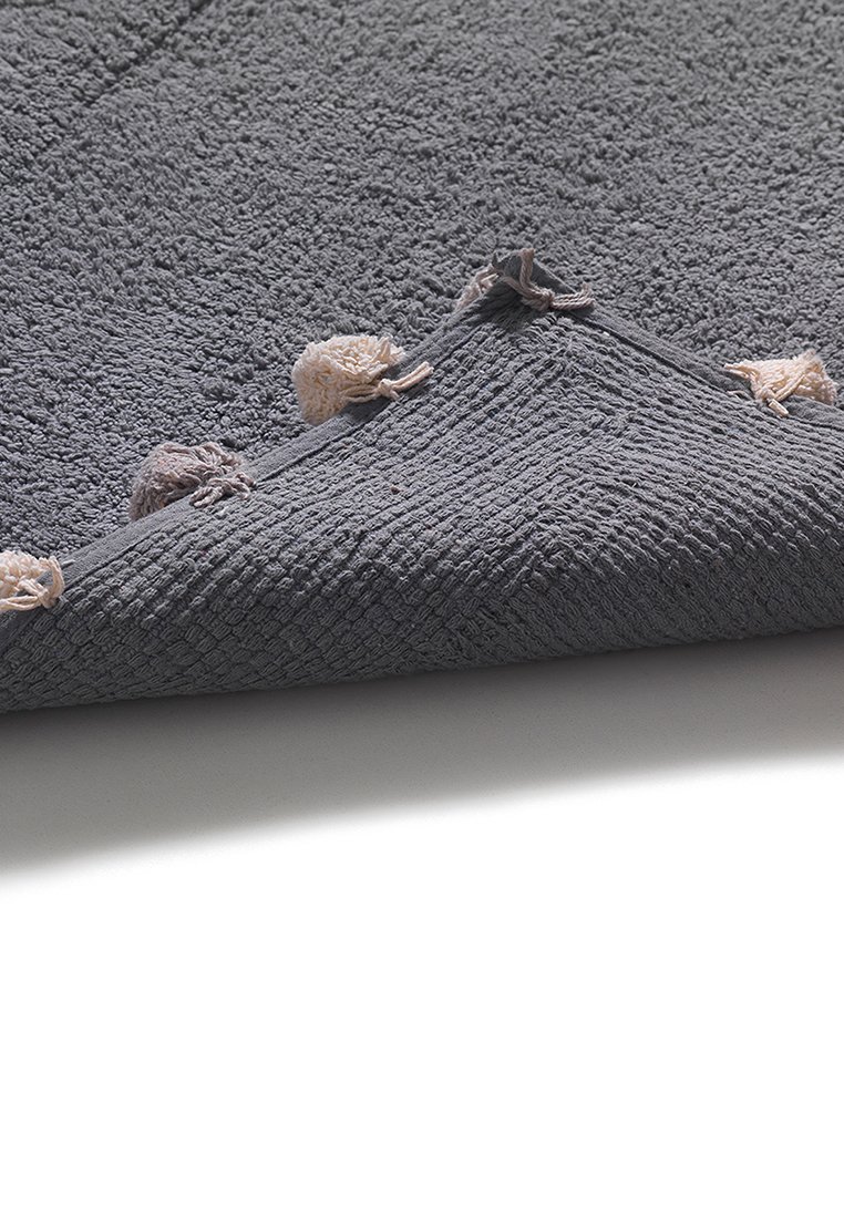 Miękki szary dywanik łazienkowy o teksturze z frędzlami, złożony w jednym rogu, ukazujący plecioną podszewkę na białej podłodze.