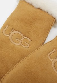 Mittens in morbido camoscio castano con fodera in pile bianco. Presentano il logo UGG ricamato in beige sulla parte anteriore. Forma curva, bordi cuciti.