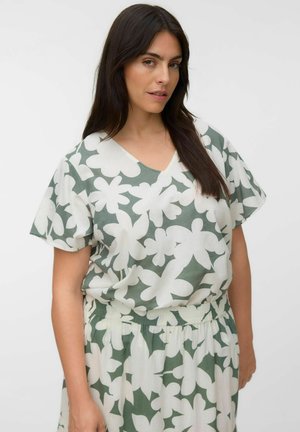 Femme aux longs cheveux foncés porte un haut court à manches vert et blanc à motif floral et une jupe assortie sur un fond clair uni.
