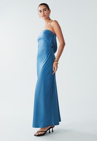Turkizna saten obleka brez naramnic z valovito silhueto, ki vsebuje stransko odprtino in subtilno draperijo, v kombinaciji s črnimi sandali s peto.