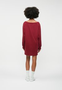 Μακρυμ άνικο, φαρδύ φόρεμα σε μπορντώ βαμβάκι, με φαρδύ ντεκολτέ και ριμπ μανσέτες, συνδυασμένο με λευκές κάλτσες και αθλητικά παπούτσια.