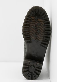 Semelle de botte noire avec des sculptures profondes, des motifs ondulés et en forme de croix, et un petit logo carré près de la zone du talon.