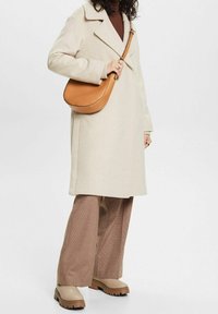 Abrigo de mezcla de lana color crema con cuello, suéter marrón, pantalones de cuadros de pierna ancha y botas beige de suela gruesa. Bolso de hombro de cuero marrón.