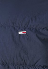 Marineblå puffjakke med en glatt tekstur, som har en liten brodert logo i rød, hvit og svart på forsiden.