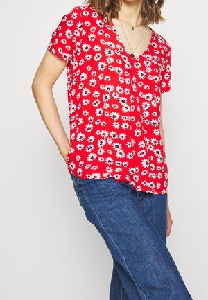 Femme portant un chemisier rouge à manches courtes avec des motifs de marguerites blanches et un jean bleu, avec une main dans la poche, sur un fond uni.