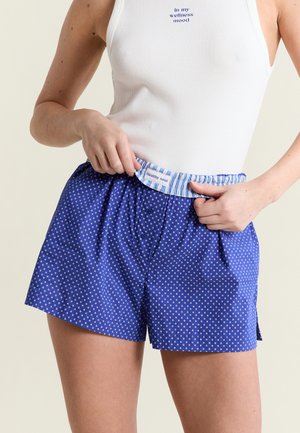 Mujer con camiseta sin mangas blanca con el texto "En mi estado de bienestar" y pantalones cortos azules con estampado y etiqueta en la cintura que dice "Alma saludable".