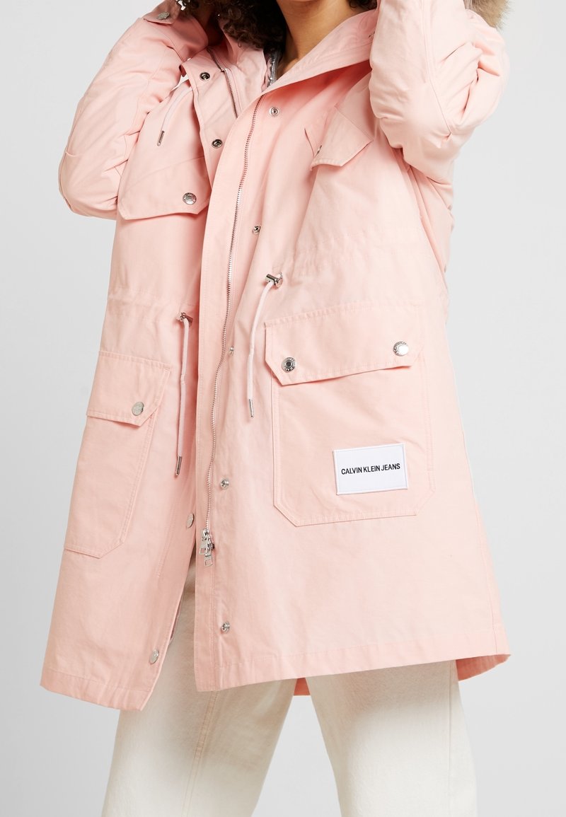 Jasnoróżowy, długi parka z kapturem, zapinany na zamek i napy, z regulowanym ściągaczem w talii, dwoma przednimi kieszeniami oraz futrzanym wykończeniem kaptura.