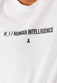 T-shirt bianco in cotone con il testo nero in grassetto "H_1/ INTELLIGENZA UMANA" e un piccolo dettaglio grafico nero sotto il testo.