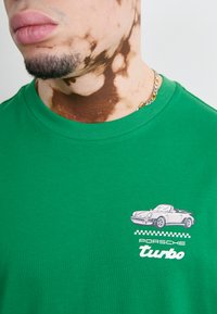 Grøn bomulds t-shirt med et brystgrafik af en vintage Porsche bil med ternede accenter og fed tekst "Porsche turbo." Casual fit.